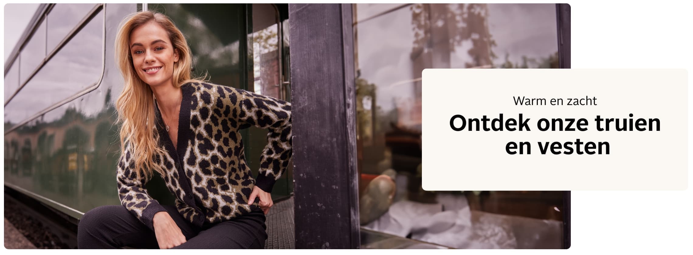 Warm en zacht - Ontdek onze truien en vesten - Vrouw in luipaardprint vest zit naast een trein